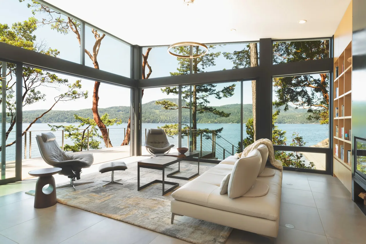 Living room of Otter Cove House features floor-to-ceiling glazing with views of water and forest setting.