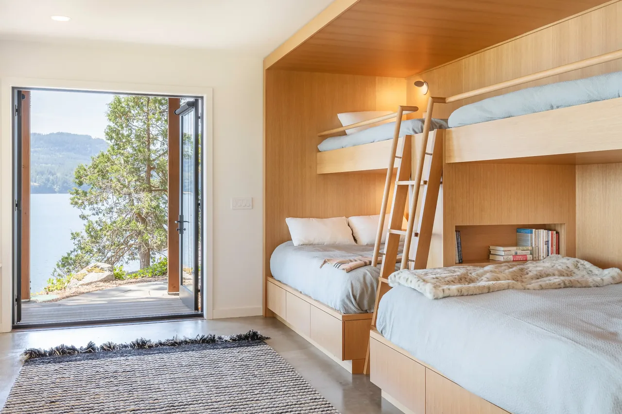 Bedroom with built-in bunk beds features light wood paneling and opens to a waterfront view.