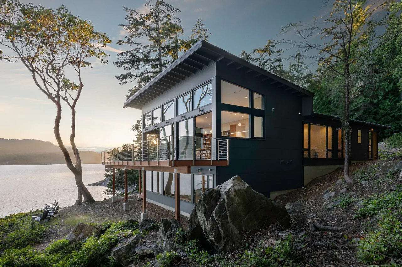 OTTER COVE HOUSE features a cantilevered balcony with metal railing and large windows overlooking a waterfront view in Seattle.