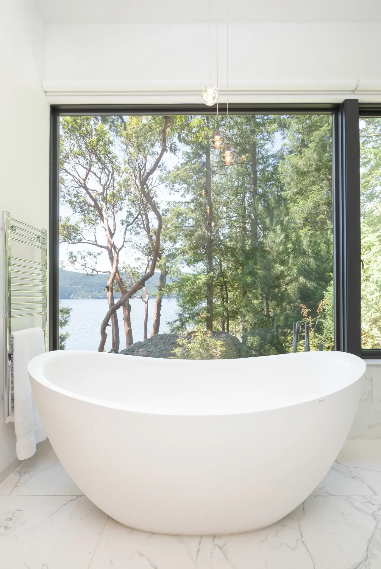 Freestanding tub in a bright bathroom with expansive views of forest and water.