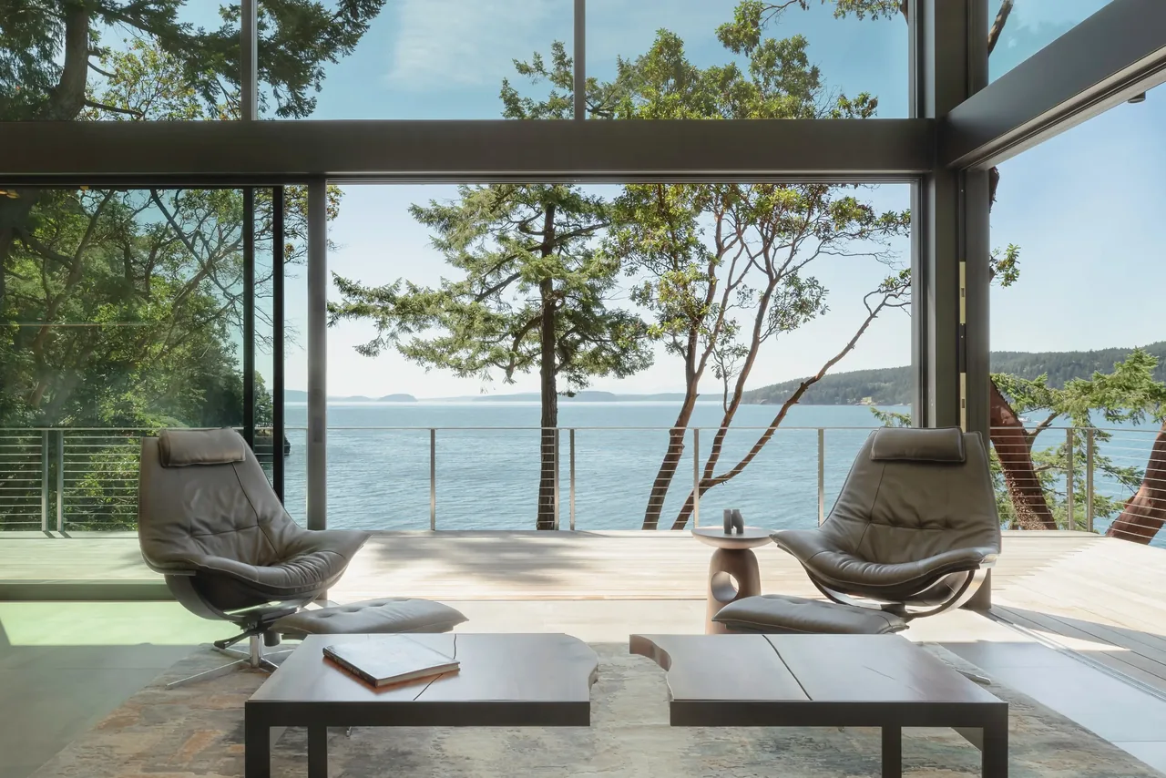 Living area of Otter Cove House with floor-to-ceiling glass walls overlooking water and trees.
