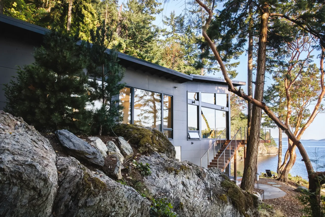 The Otter Cove House features large windows and a metal-railed exterior stair, blending into a rocky forest site by the water.