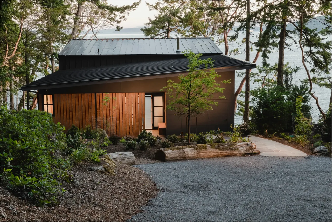 OTTER COVE HOUSE by Stoltz Kau Architects features clean lines, a black metal roof, and wood siding, set in a forested site near the water.