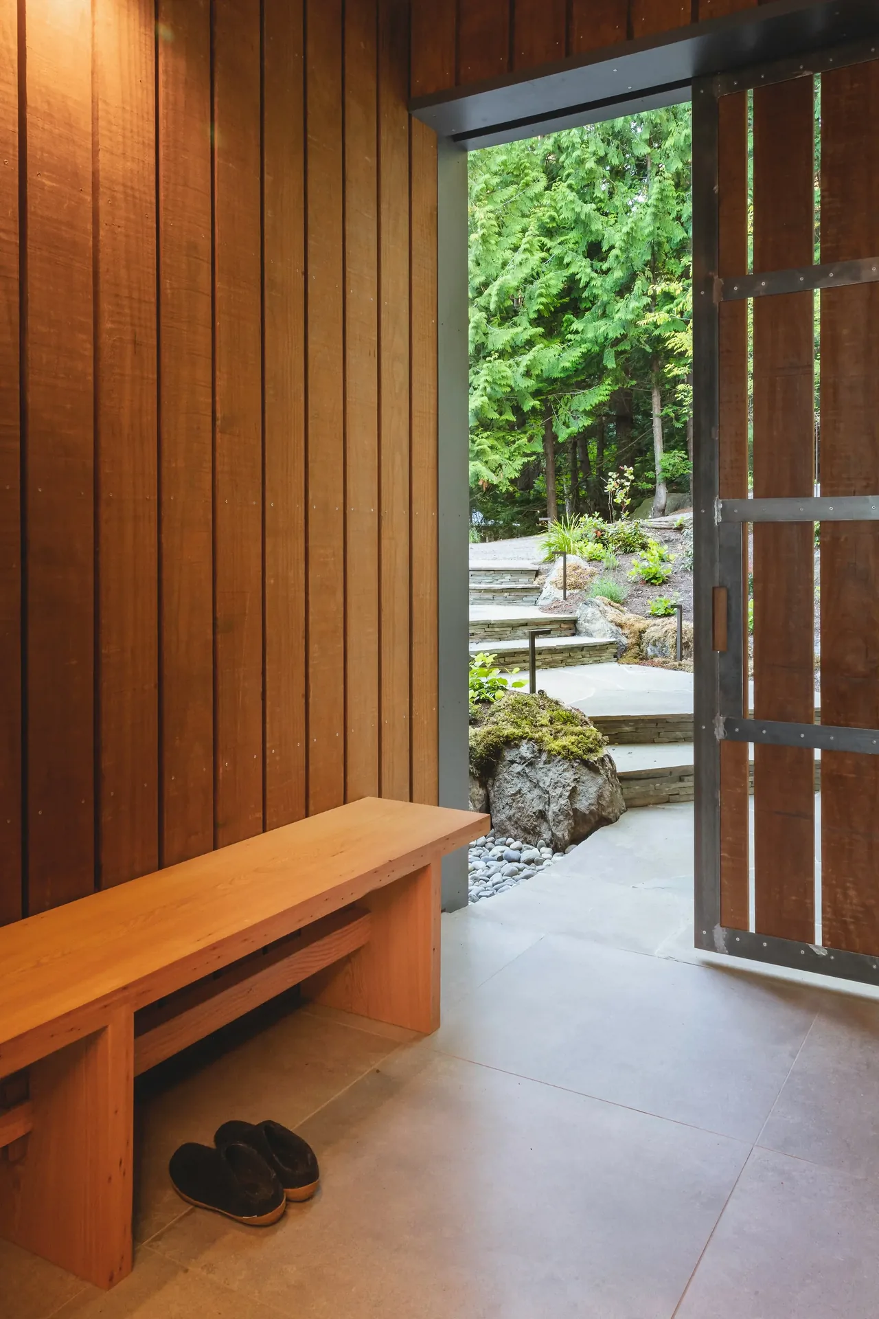 The entryway features vertical wood paneling, a simple wooden bench, and opens to a landscaped stone stair outside.