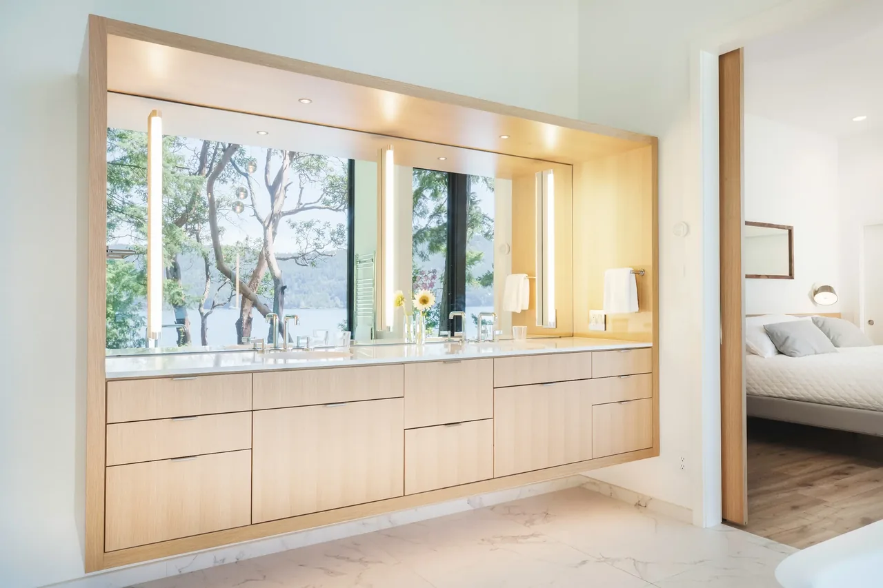 Bathroom vanity with dual sinks and built-in lighting, featuring a large mirror reflecting a waterfront and forest scene.
