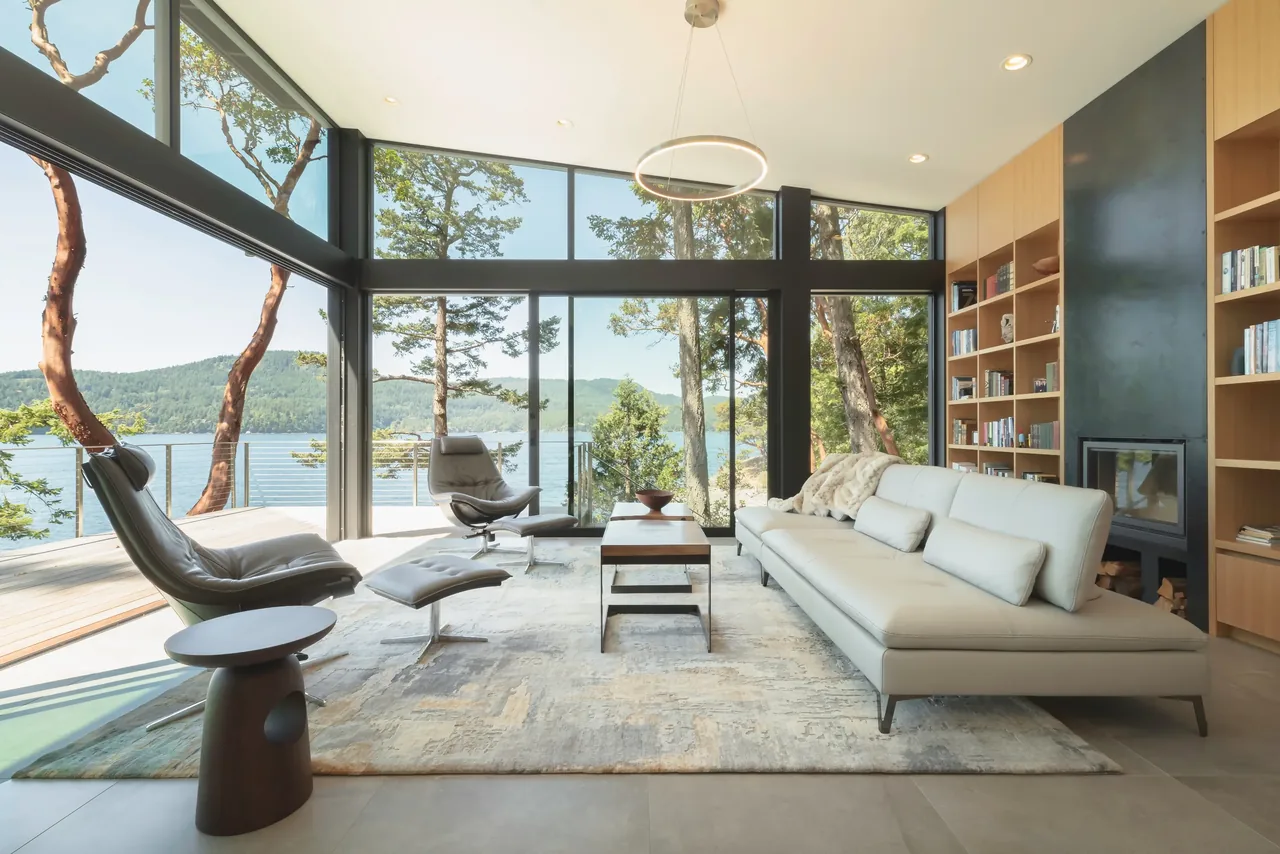 Living room with floor-to-ceiling glazing offers expansive lake and forest views, furnished with modern leather seating and a built-in bookcase with fireplace.