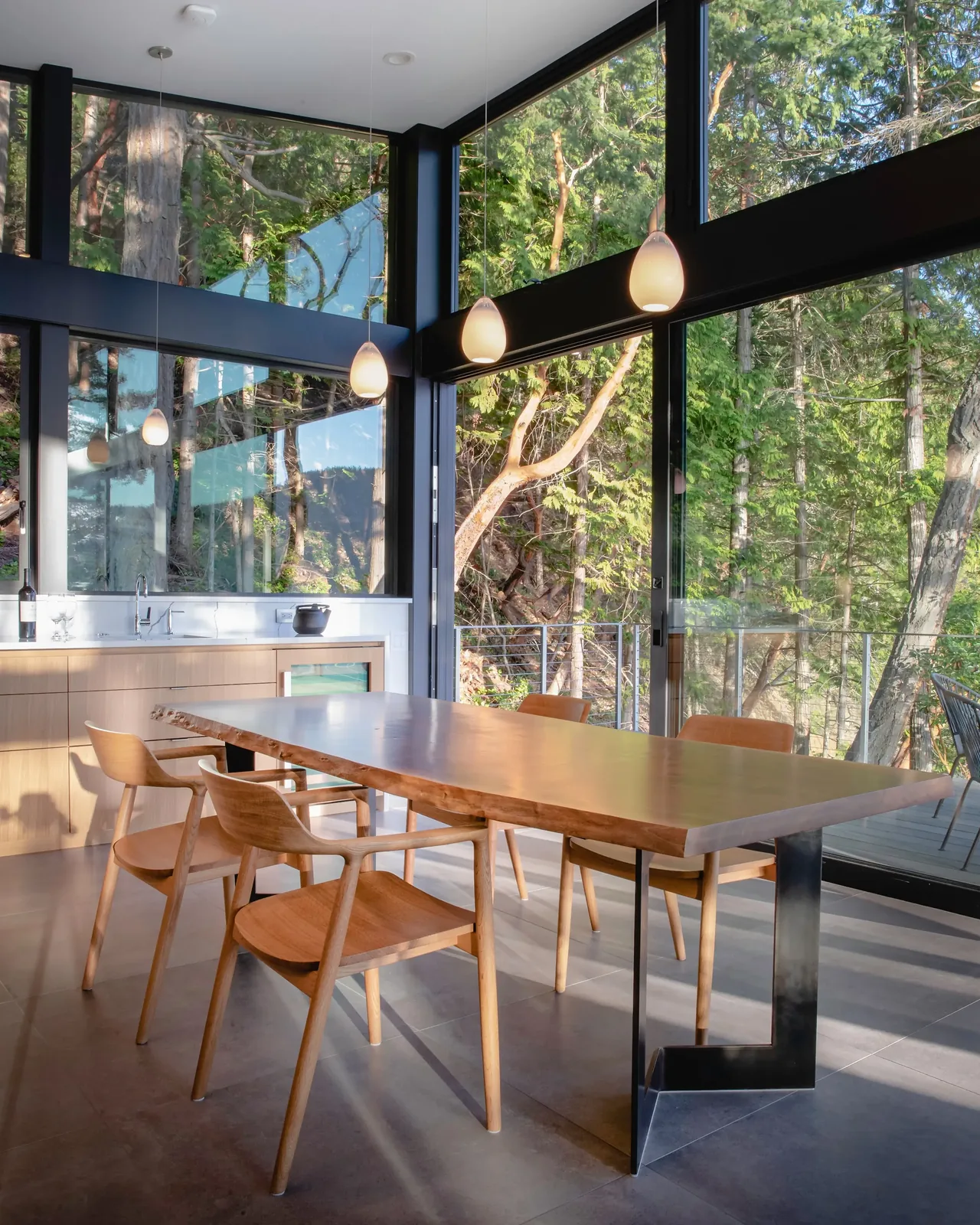 Dining room features a live-edge wooden table, wood chairs, and floor-to-ceiling glass windows overlooking a forested landscape.