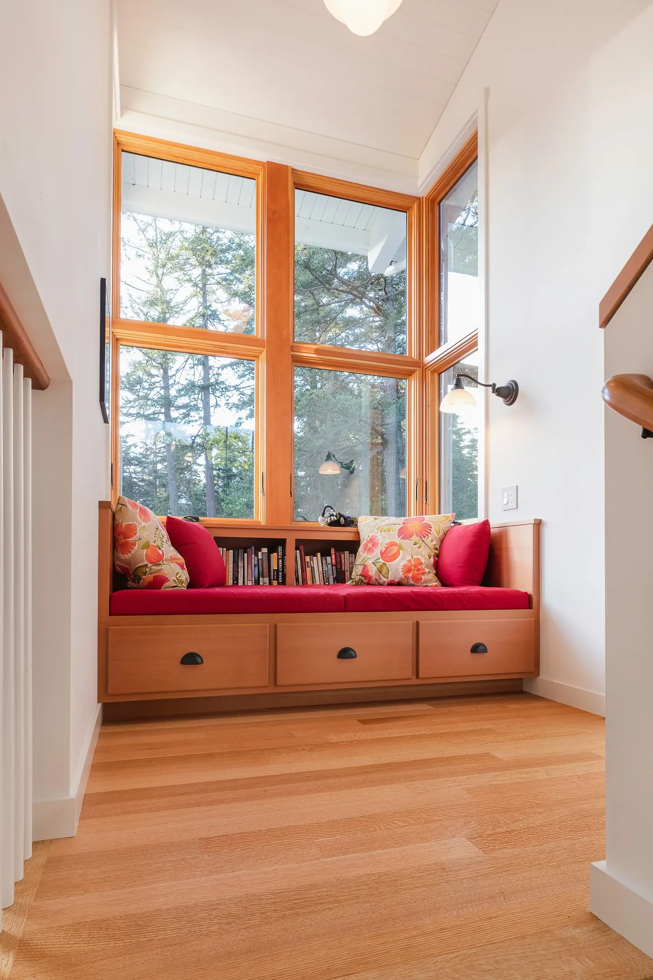 Built-in wooden window seat with storage drawers, red cushions, and floral pillows under tall wood-framed windows.