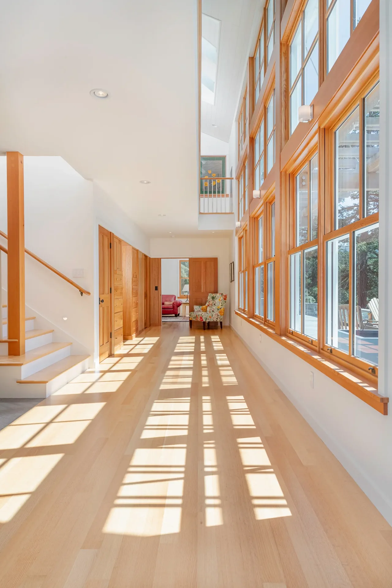A hallway featuring large wood-framed windows and light wood flooring with wood-paneled doors and a staircase.