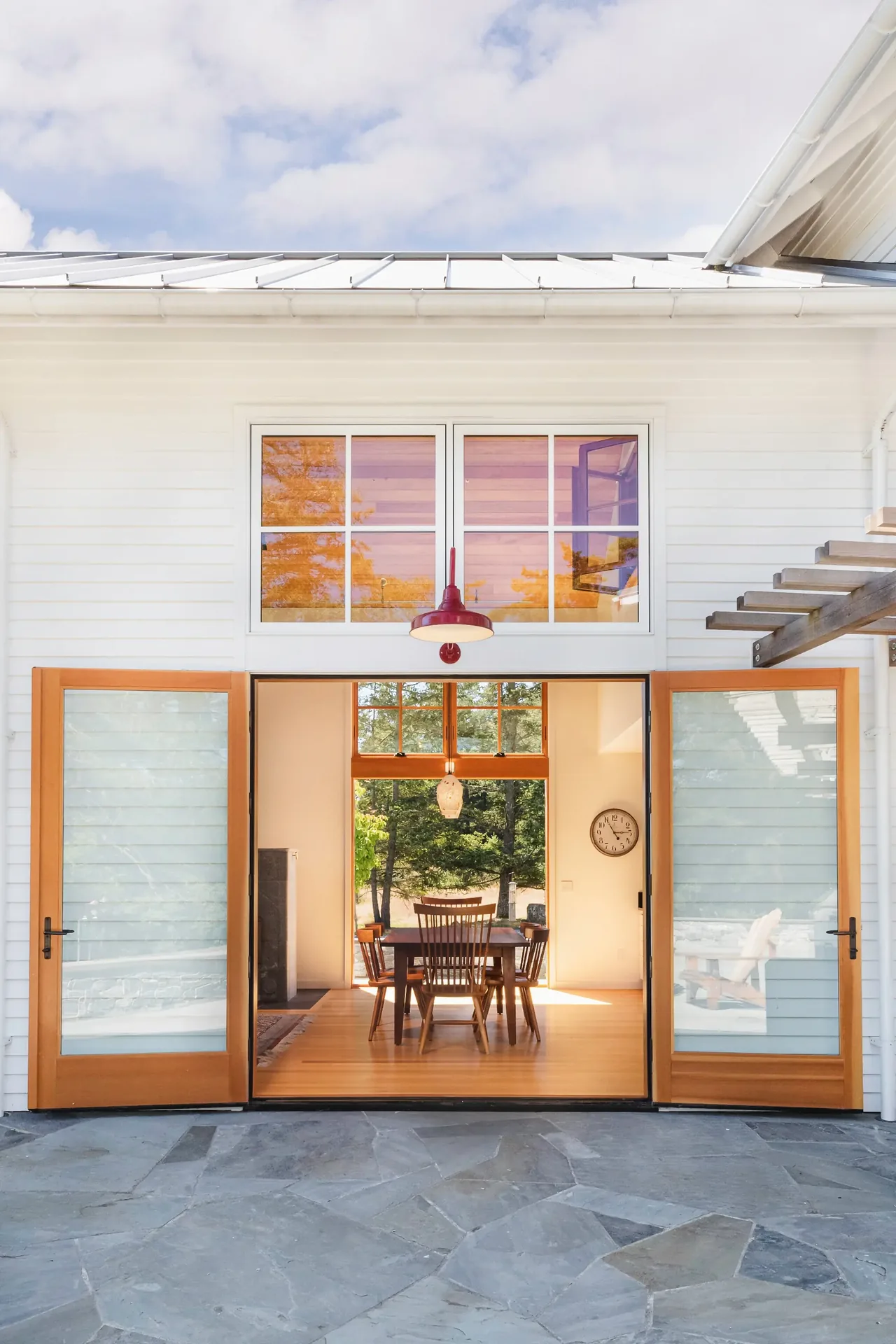 Open wood and glass double doors lead from stone patio into a dining room with wood furniture and a large window.