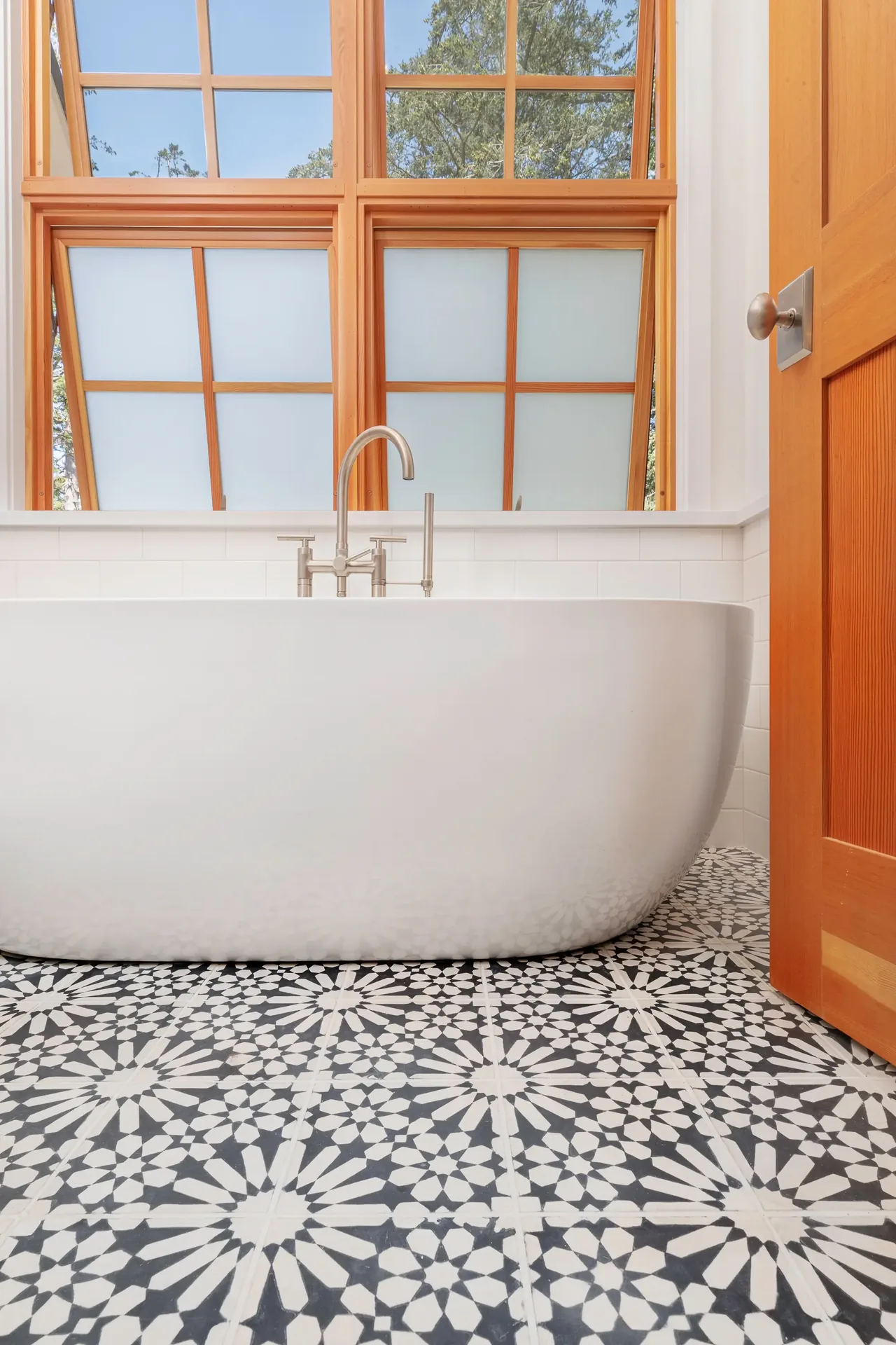 Bathroom with a white freestanding tub, patterned black and white floor tiles, and wood-framed paneled windows.