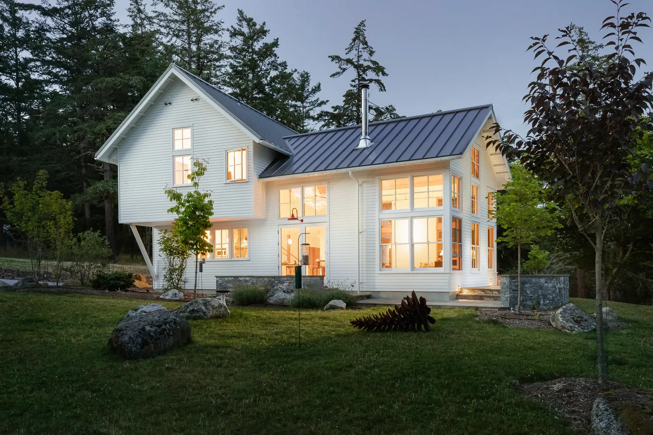 MAP CORNER, a home featuring white wood siding and standing seam metal roofing, with illuminated large windows among trees.