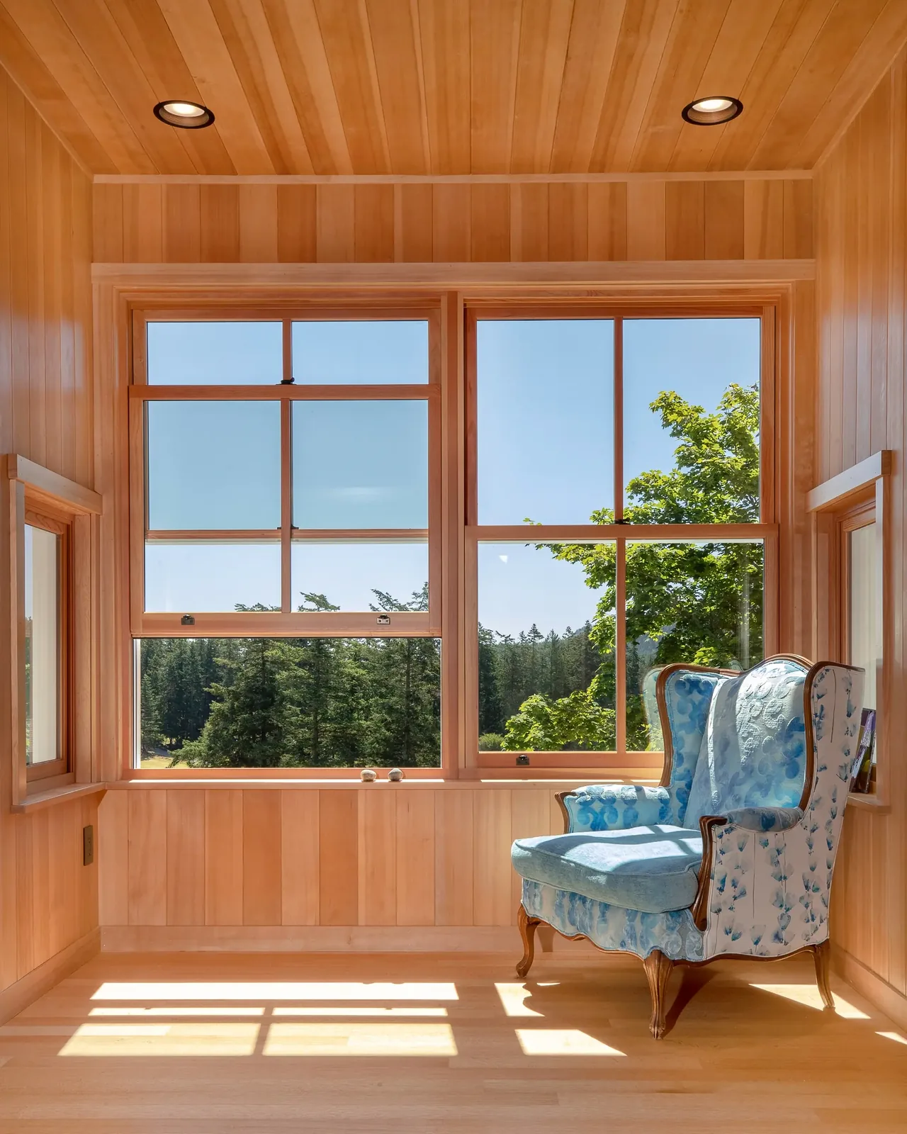 Wood-paneled corner room with large windows overlooks a wooded landscape, furnished with a blue wingback armchair.
