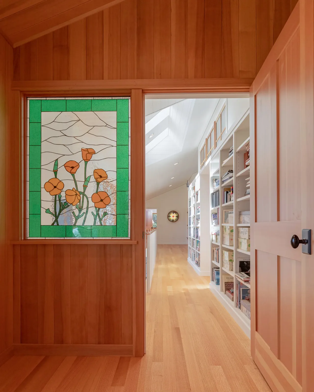Hallway with built-in white bookshelves, stained glass floral window, and natural wood paneling and flooring.