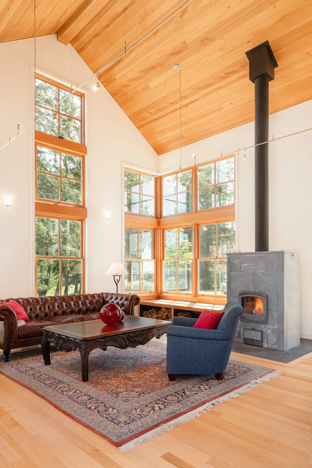 Living room with vaulted wood ceiling and multi-pane windows overlooking greenery, featuring a stone fireplace and seating area.