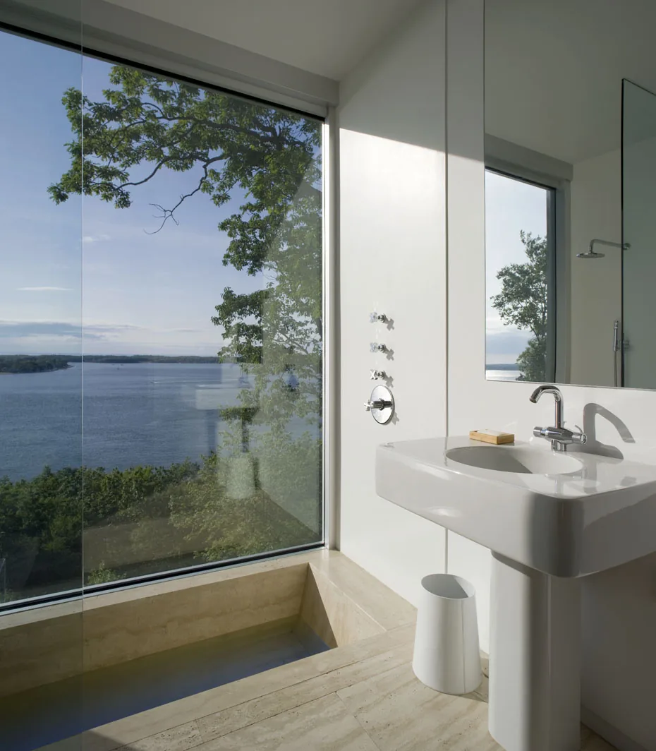 Bathroom features a large glass window with a water view, a beige tiled floor, and a white pedestal sink.