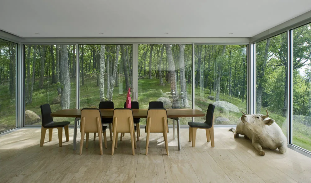 Clearhouse dining area features a wooden table with mixed chairs, enclosed by floor-to-ceiling windows overlooking trees and nature.