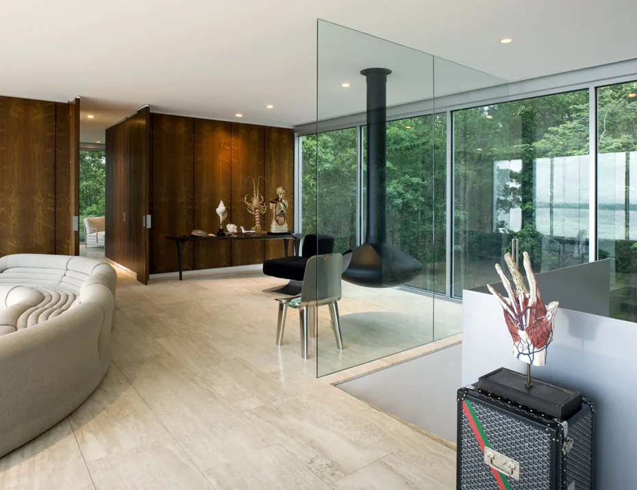 Clearhouse living room features floor-to-ceiling glazing with views of trees and water, highlighted by a suspended black fireplace and sculptural decor.
