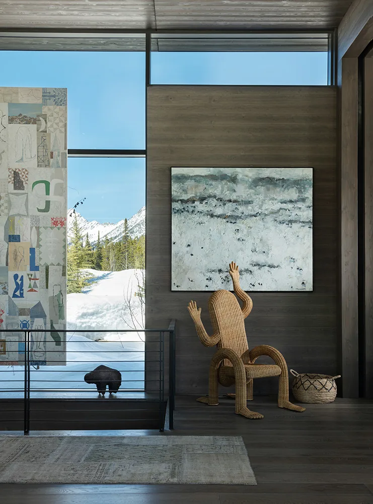 Living area shows art-filled wall, wicker human-shaped chair, and a large window with snow-covered trees outside.