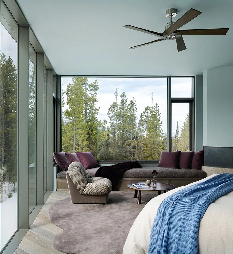 Bedroom at Moonlight Basin project with expansive windows and forest views, featuring a cushioned window bench and patterned armchair.