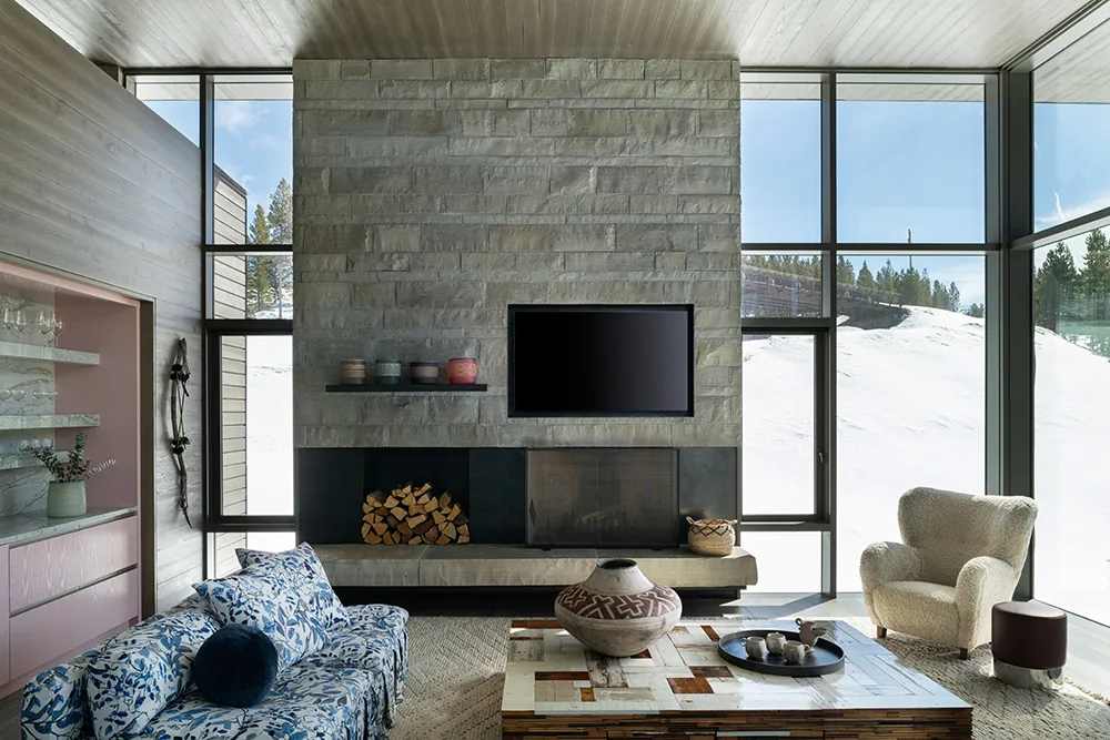 The living room features a stone fireplace wall with a mounted TV and floor-to-ceiling windows overlooking a snowy landscape in Moonlight Basin.
