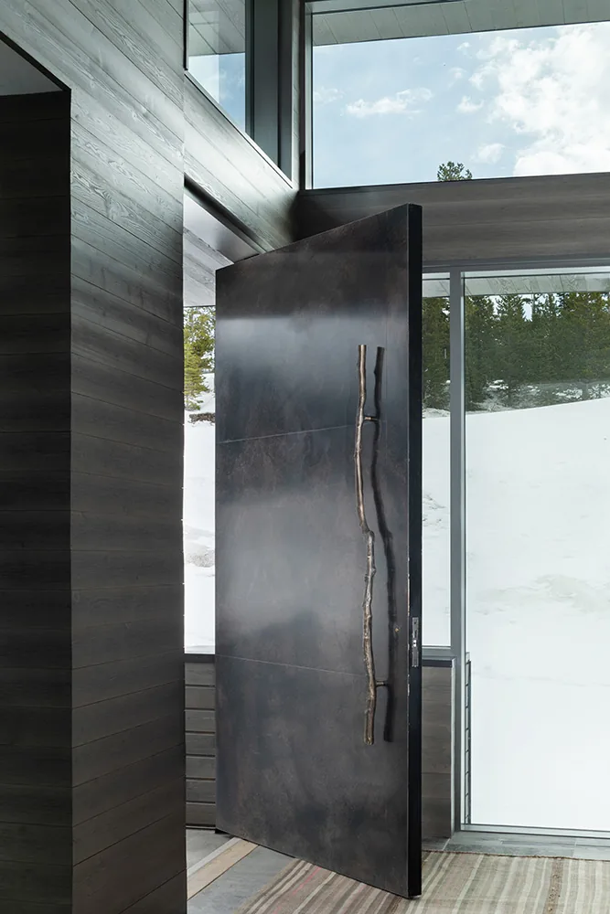 Pivot door with a natural branch handle opens to a snow-covered landscape visible through floor-to-ceiling windows.