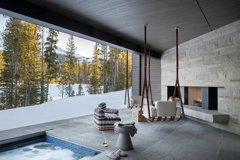 Covered patio area featuring suspended chairs, a stone fireplace, and a pool with snow and forest beyond.
