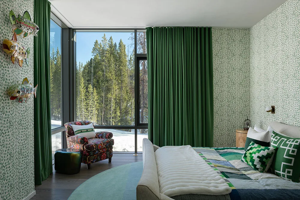 Bedroom featuring green patterned wallpaper, large corner windows with green curtains, and a colorful armchair next to a snowy forest view.