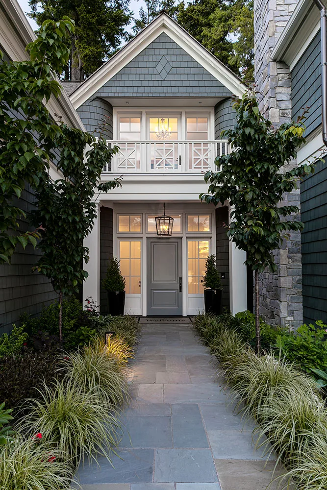 Tangled Ride house entry features gray shingle siding, white trim, multiple gables, and stone chimney with landscaped stone pathway.