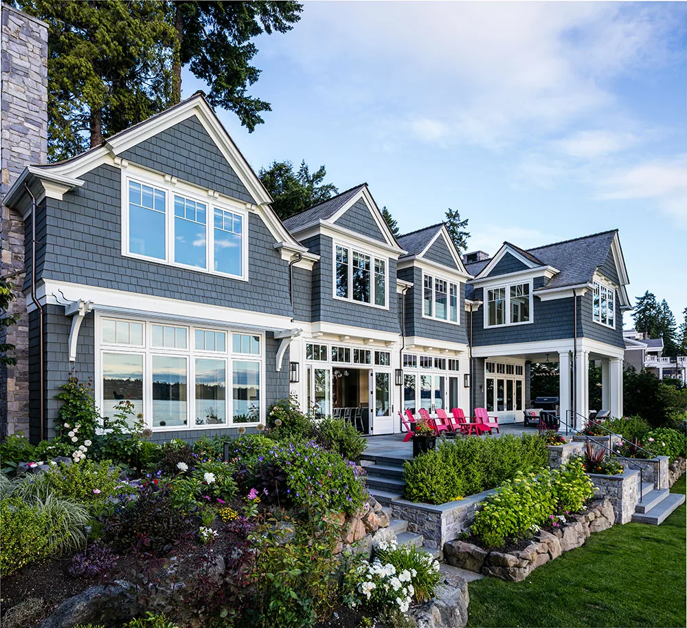 The 'Tangled Ride' residence features gray shingle siding, multiple gable roofs, and large windows overlooking a landscaped garden and lake.