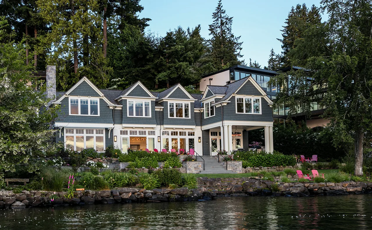 A lakeside two-story house with shingle siding, multiple gable roofs, and landscaped stone terraces leading to the water's edge.