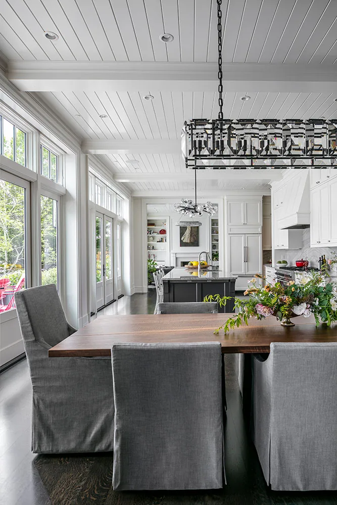 Open dining area with gray upholstered chairs and dark wood table connects to a white kitchen with an island and large windows.