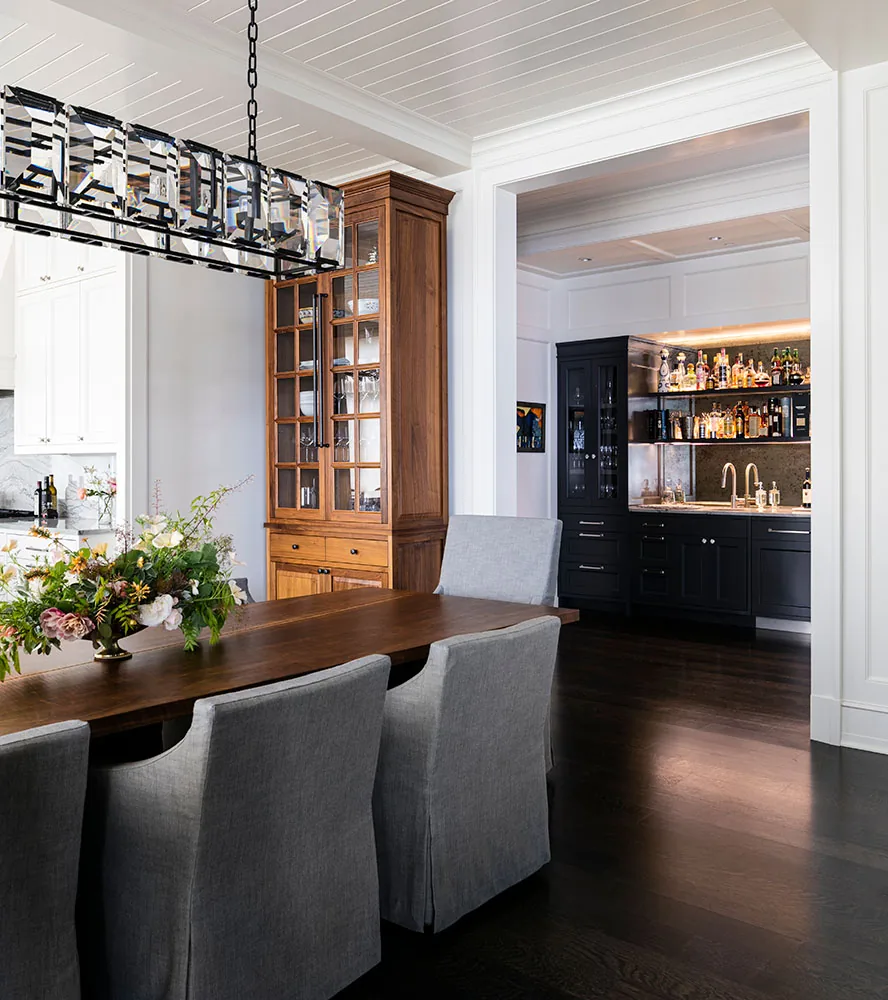 Dining room with gray upholstered chairs and a wood cabinet, looking into a bar area with black cabinets and a sink.
