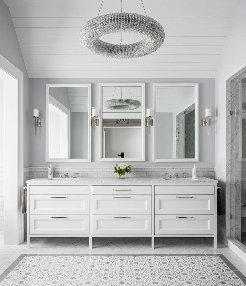 A white double vanity with marble countertop is topped by three mirrors and flanked by wall sconces beneath a circular crystal chandelier.