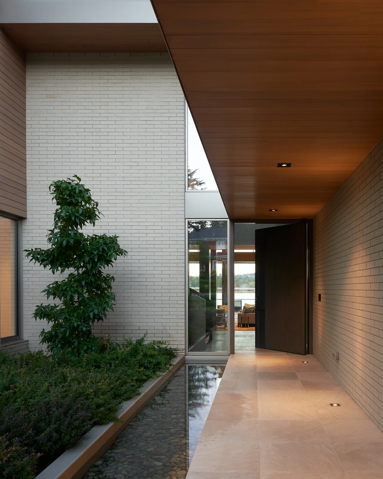 Covered entry of Union Bay Residence featuring wood-paneled ceiling and stone tile floor with adjacent planted garden and water feature.