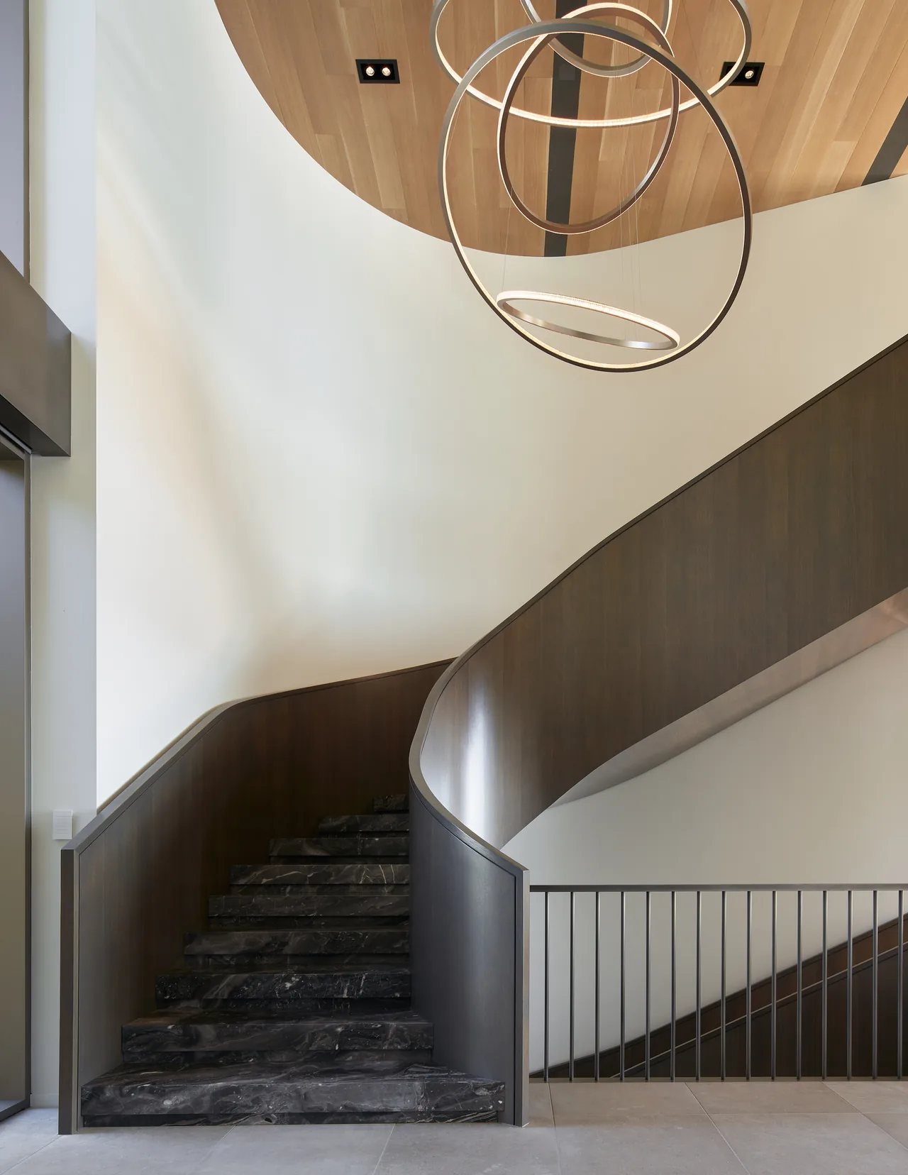A curved interior staircase features black marble steps, dark wood railing, and modern ring pendant lighting beneath a circular wood ceiling feature.