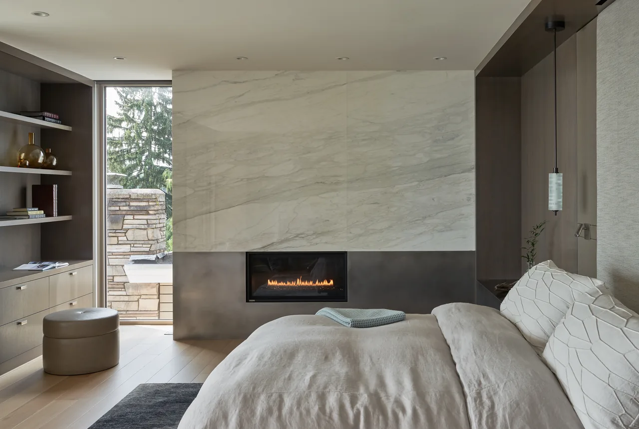 Bedroom features a linear fireplace beneath a large marble panel wall and built-in shelving with natural wood tones.