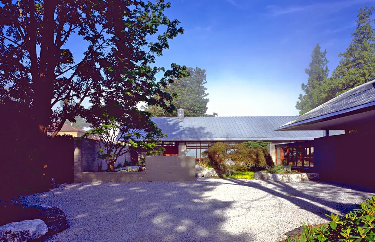 Solbeck Residence features a gravel driveway and a curved metal roof, set among mature trees and landscaping.