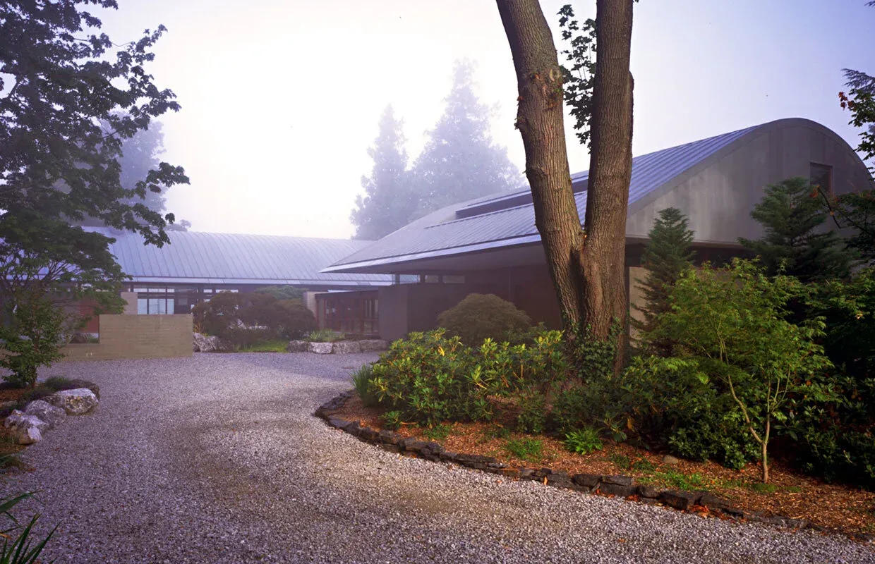 Exterior view of Solbeck Residence with curved metal roof and gravel driveway surrounded by trees and shrubs in foggy weather.