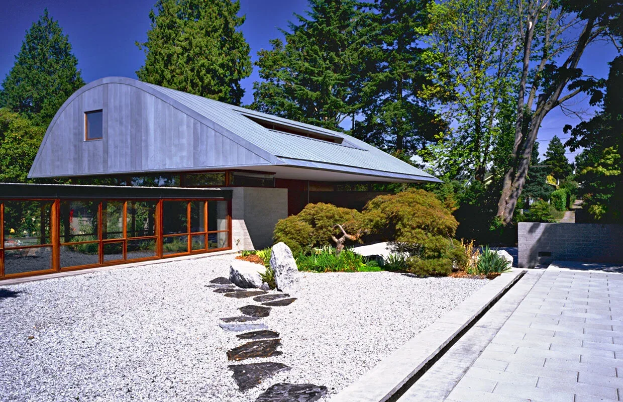 The Solbeck Residence features a distinct curved metal roof, wood-framed large windows, and a gravel driveway, nestled among lush landscaping.