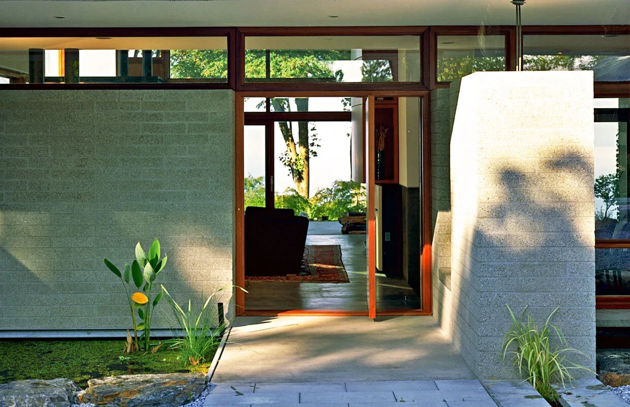 Exterior front of Solbeck Residence showing a wooden frame glass front door and adjacent stone walls with greenery.