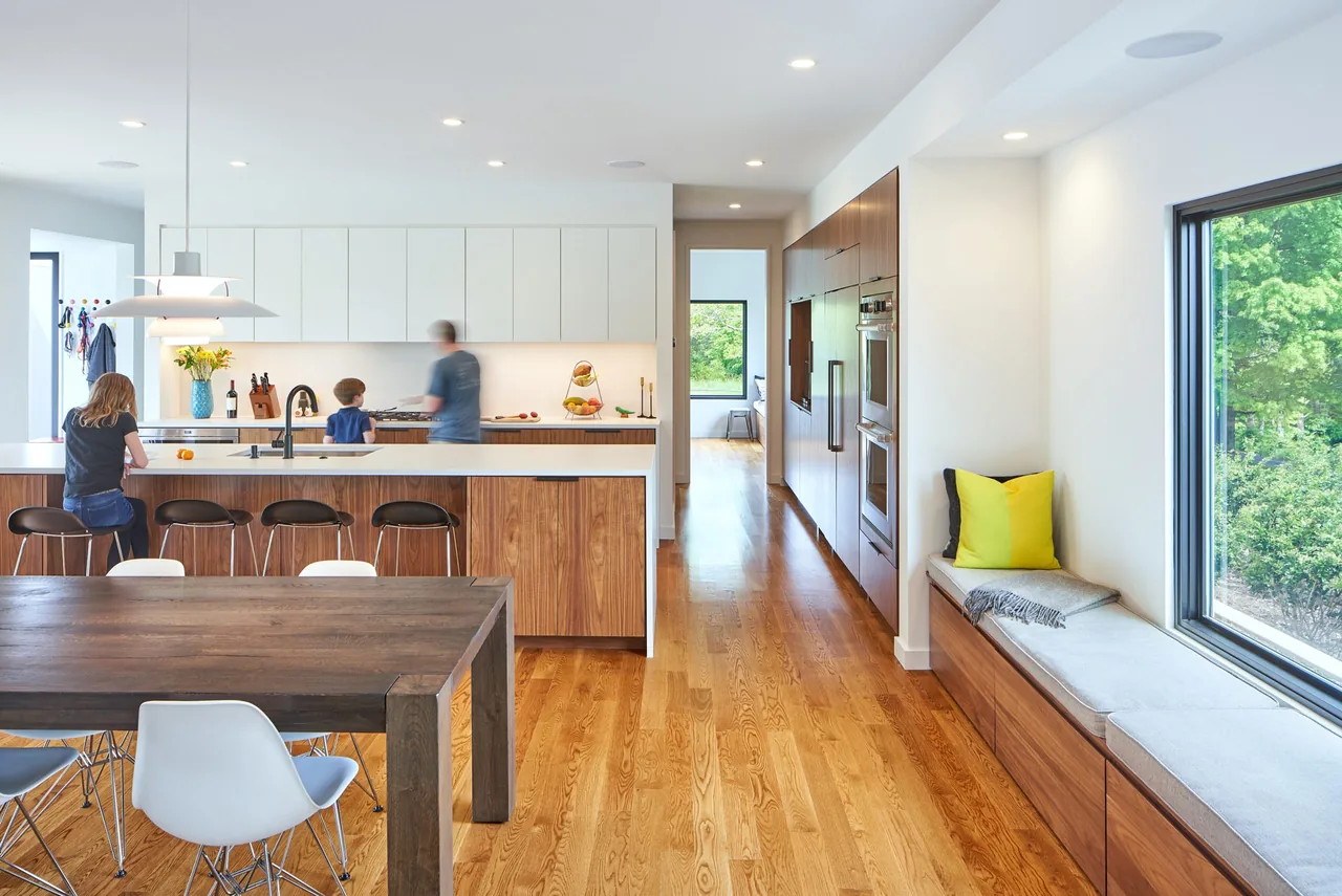 Open kitchen with white upper cabinets and wood lower cabinets, adjacent to a dining table and window seat with colorful cushions.