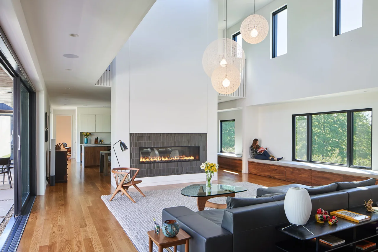 Living room features a double-height ceiling, wood flooring, and large windows with black frames and built-in bench seating.