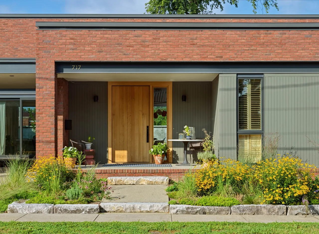 The entrance of Morgan Street House features a wooden door set in green siding with a brick facade above and flower beds in front.