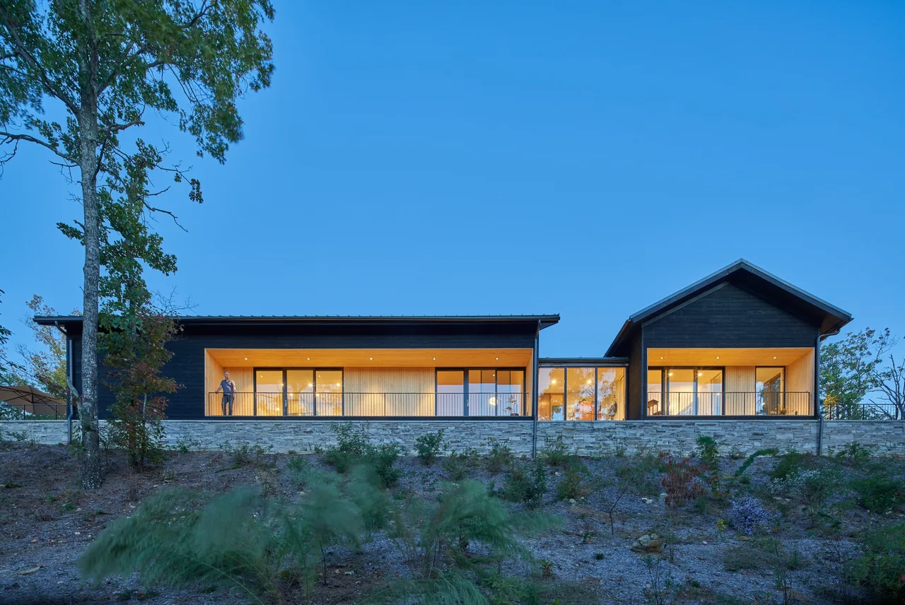 Short Mountain House features connected volumes with recessed balconies illuminated against a stone retaining wall.