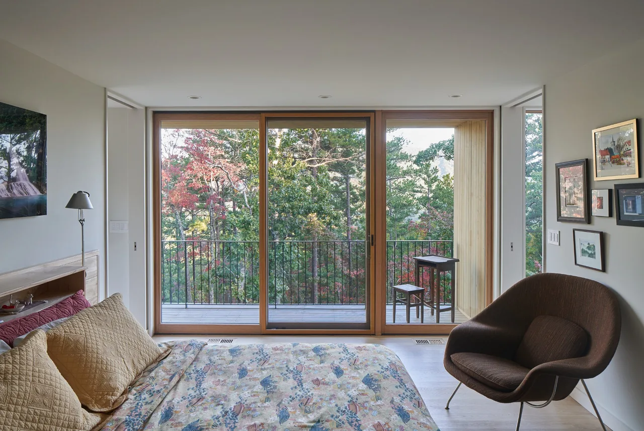 This bedroom features large wood-framed sliding doors opening to a balcony with forest views in fall colors.