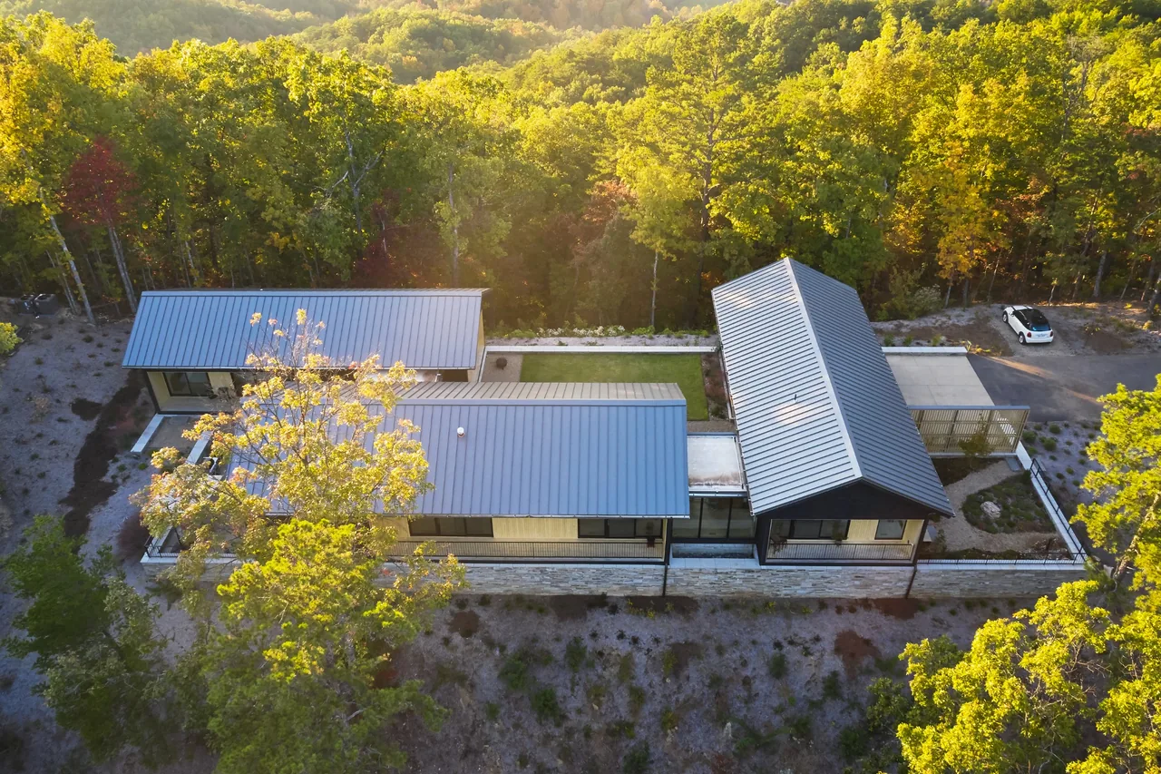 Short Mountain House features three connected volumes with standing seam metal roofs and large glass walls, set in a wooded area.