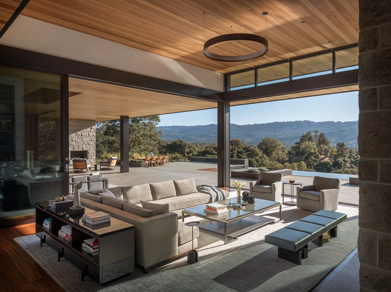Open living room with modern furniture and large glass walls overlooking patio and mountain landscape.