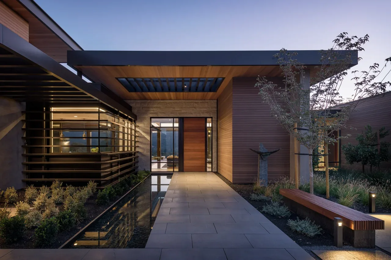 Portola Valley House entry features wood siding, a stone wall, water feature, and pathway lighting at dusk.