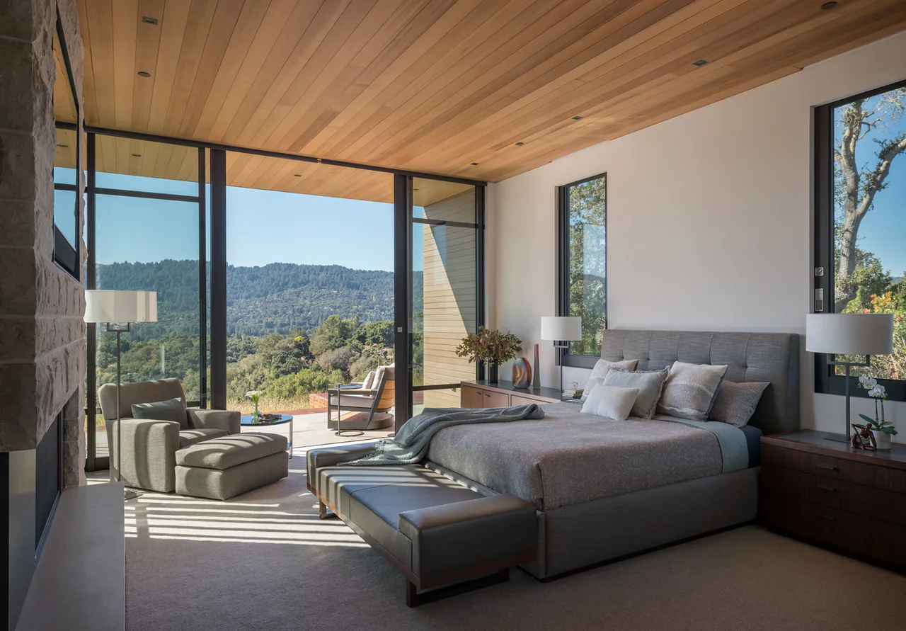Bedroom with floor-to-ceiling glazing revealing mountain views, wood ceiling, and stone fireplace.
